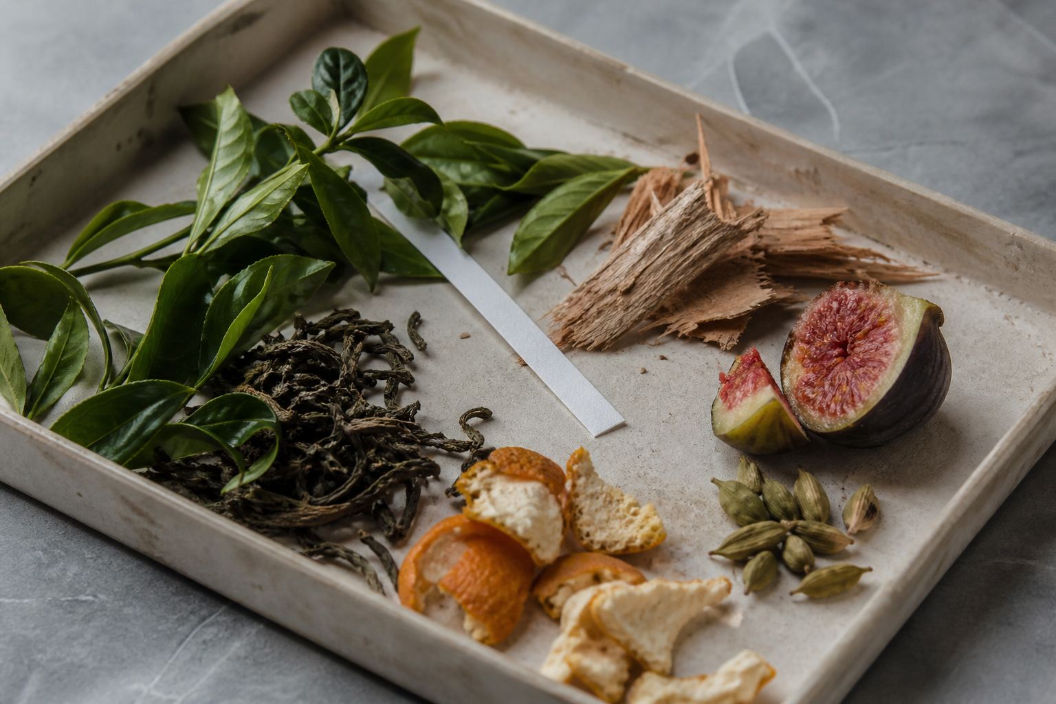 Green tea leaves, dried tea, fig, cedar, orange peel and cardamom arranged on an archival tray