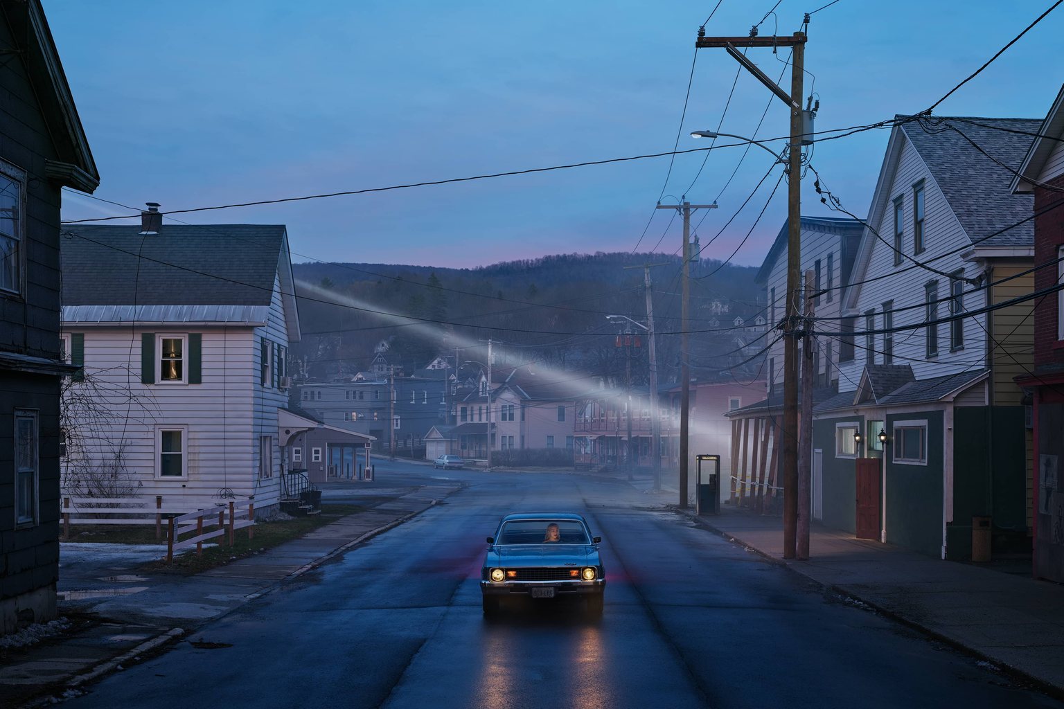 Phoebe Bridgers seated in a blue car on an empty misty small-town street at dusk