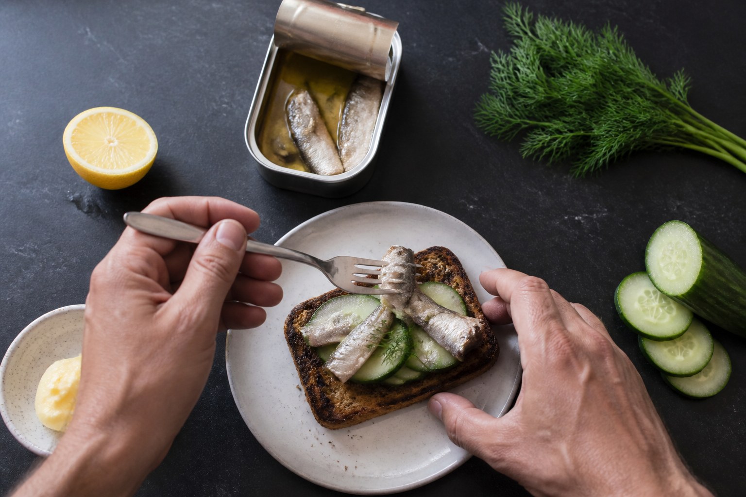 Two hands using a fork to place sardines over cucumber and grilled rye toast on a dark kitchen counter