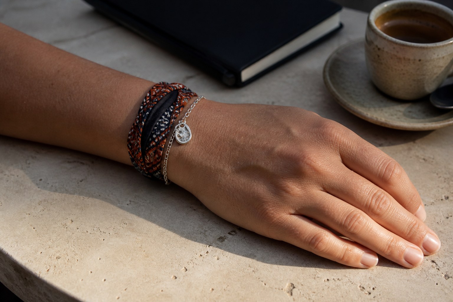 Rust-and-ink silk bandana bracelet wrapped with a fine silver chain and small abstract charm on a stone café table