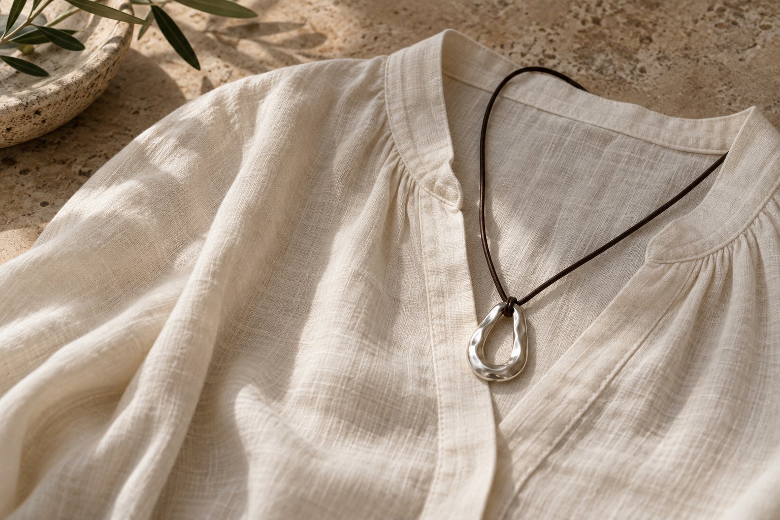 Sculptural silver pendant on a dark leather cord draped over a cream linen blouse on limestone