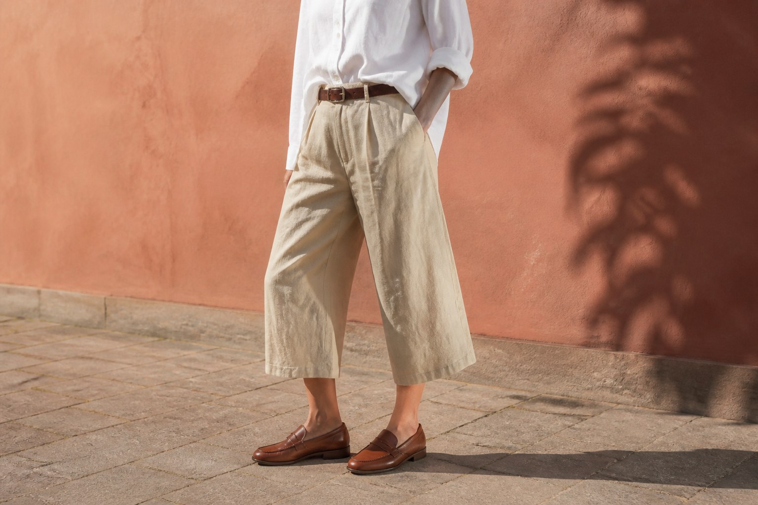 Sand linen-blend wide capri trousers with a white shirt, slim brown belt and tobacco loafers against terracotta plaster