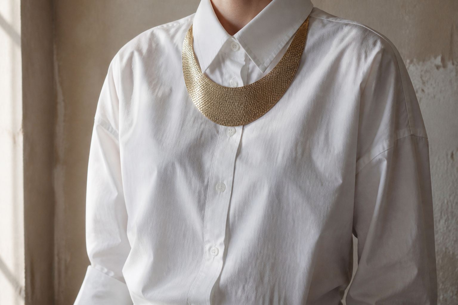 Cropped white cotton shirt styled with a broad hammered gold bib necklace, with no face visible