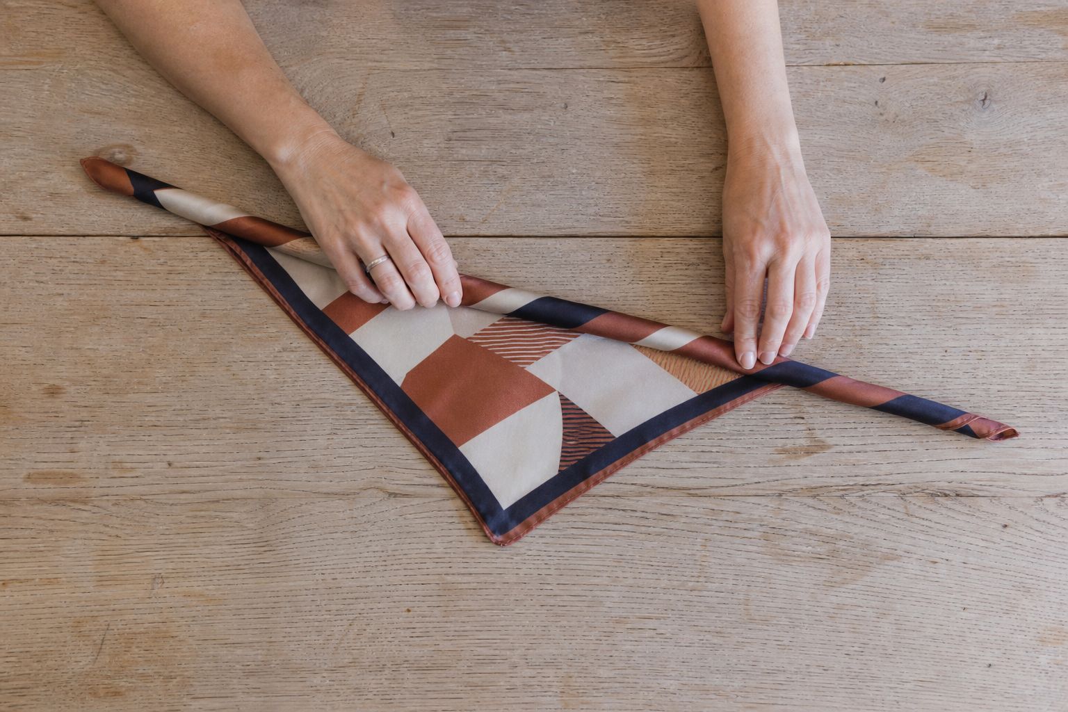 Hands folding a burgundy, cream and olive silk scarf diagonally on an oak table