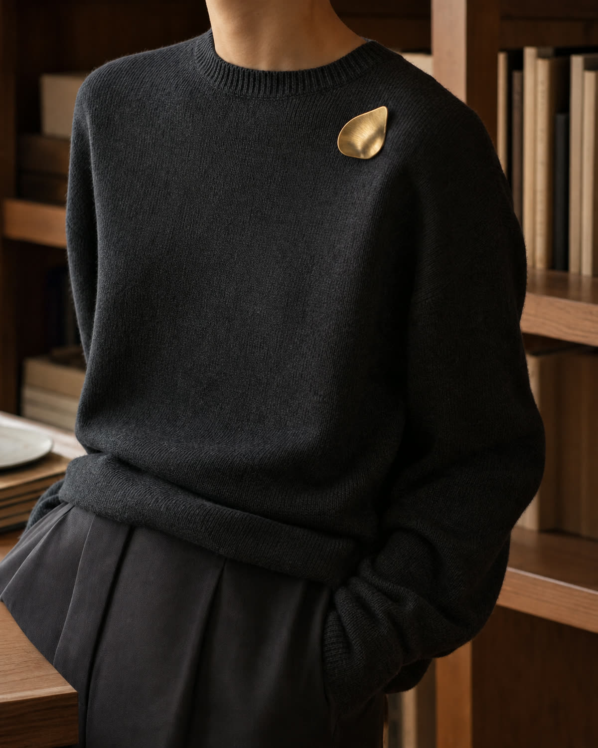 Brushed-gold sculptural brooch pinned high on the shoulder of a dense charcoal jumper