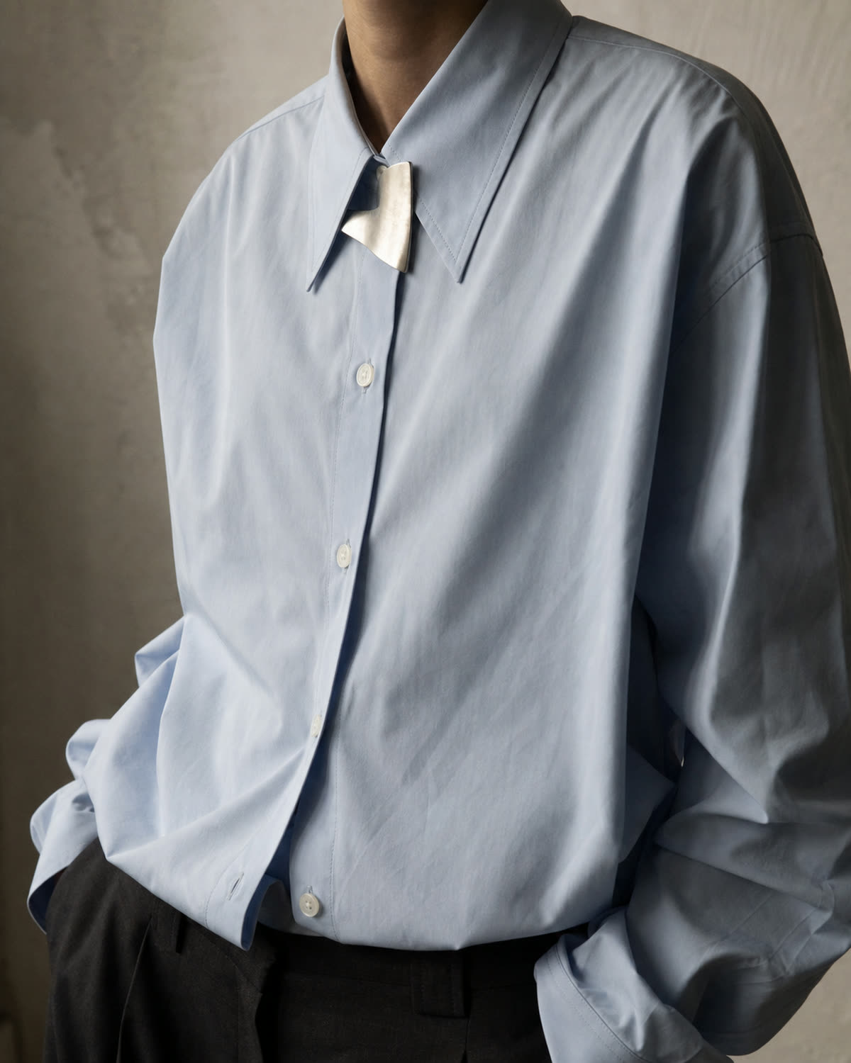 Small brushed-silver brooch pinned between the collar points of an oversized pale-blue shirt