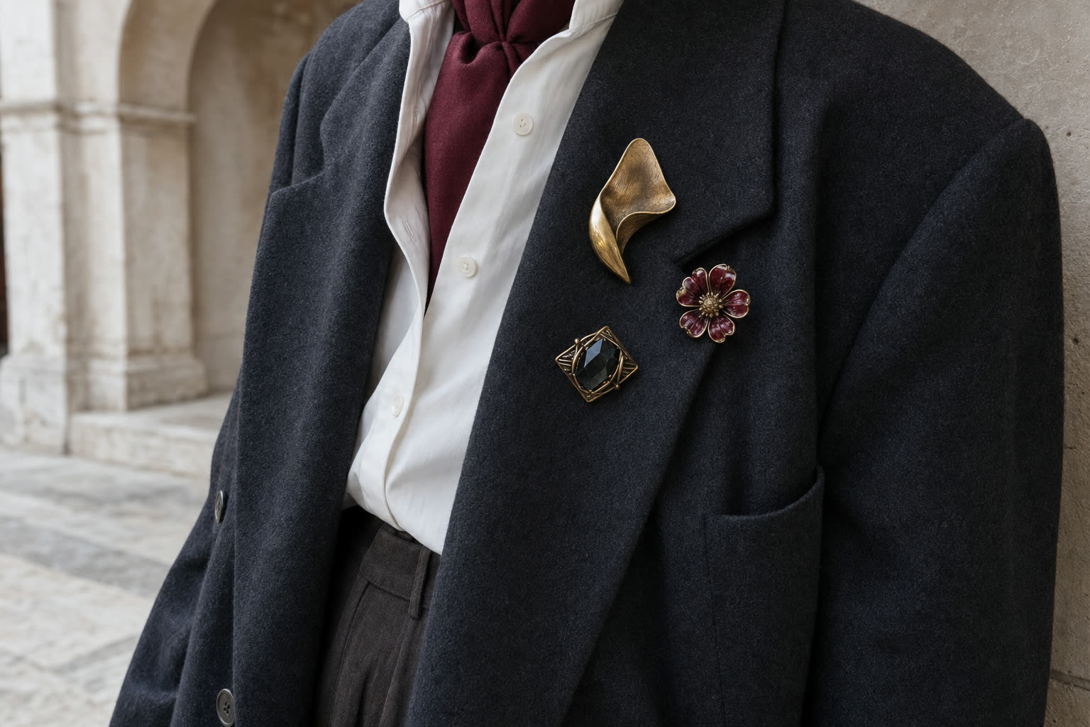 Three antique-style brooches arranged on an oversized charcoal blazer with an oxblood scarf
