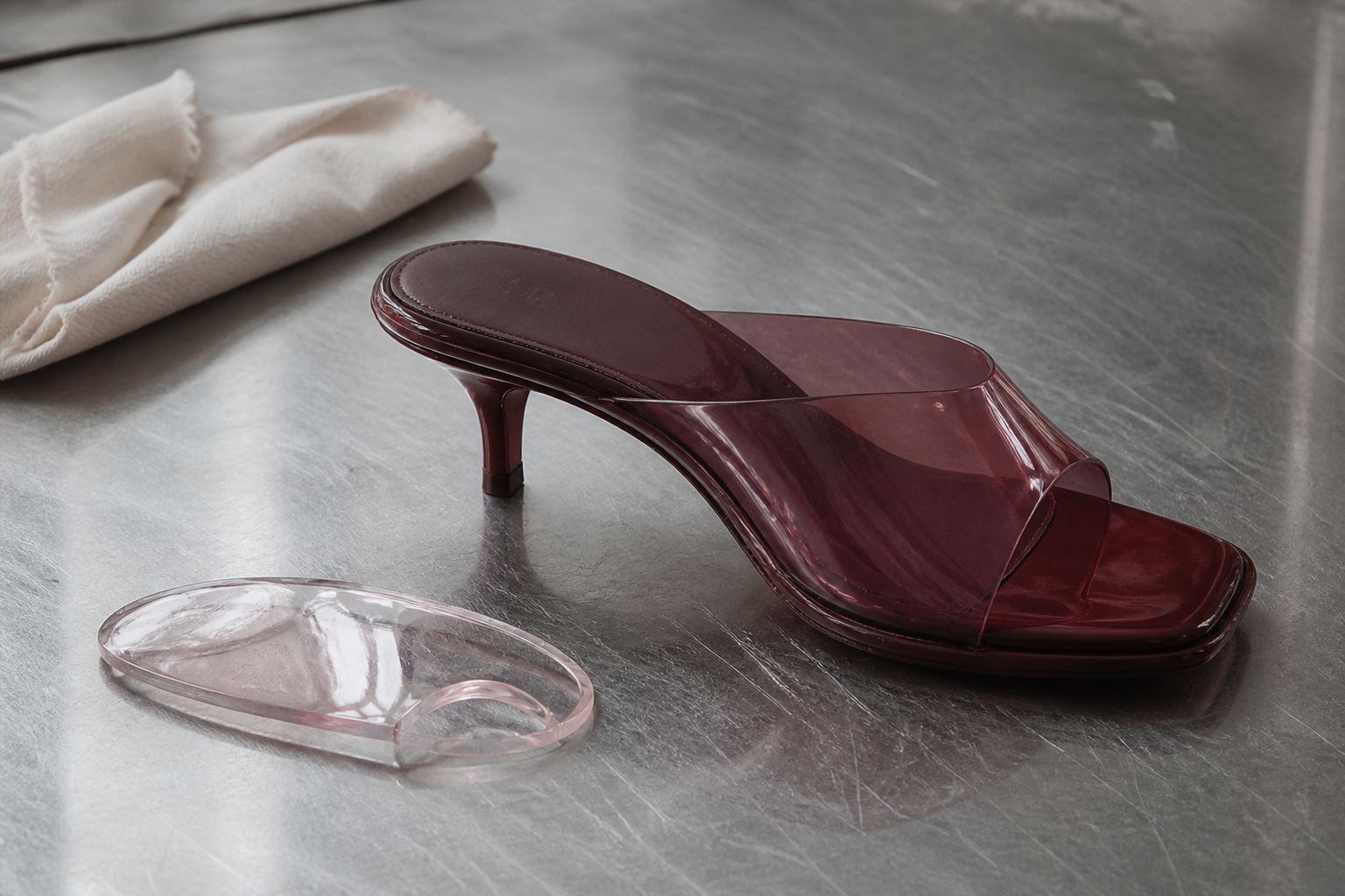 Translucent oxblood jelly kitten-heel mule on a scratched steel worktable showing its moulded upper and low heel