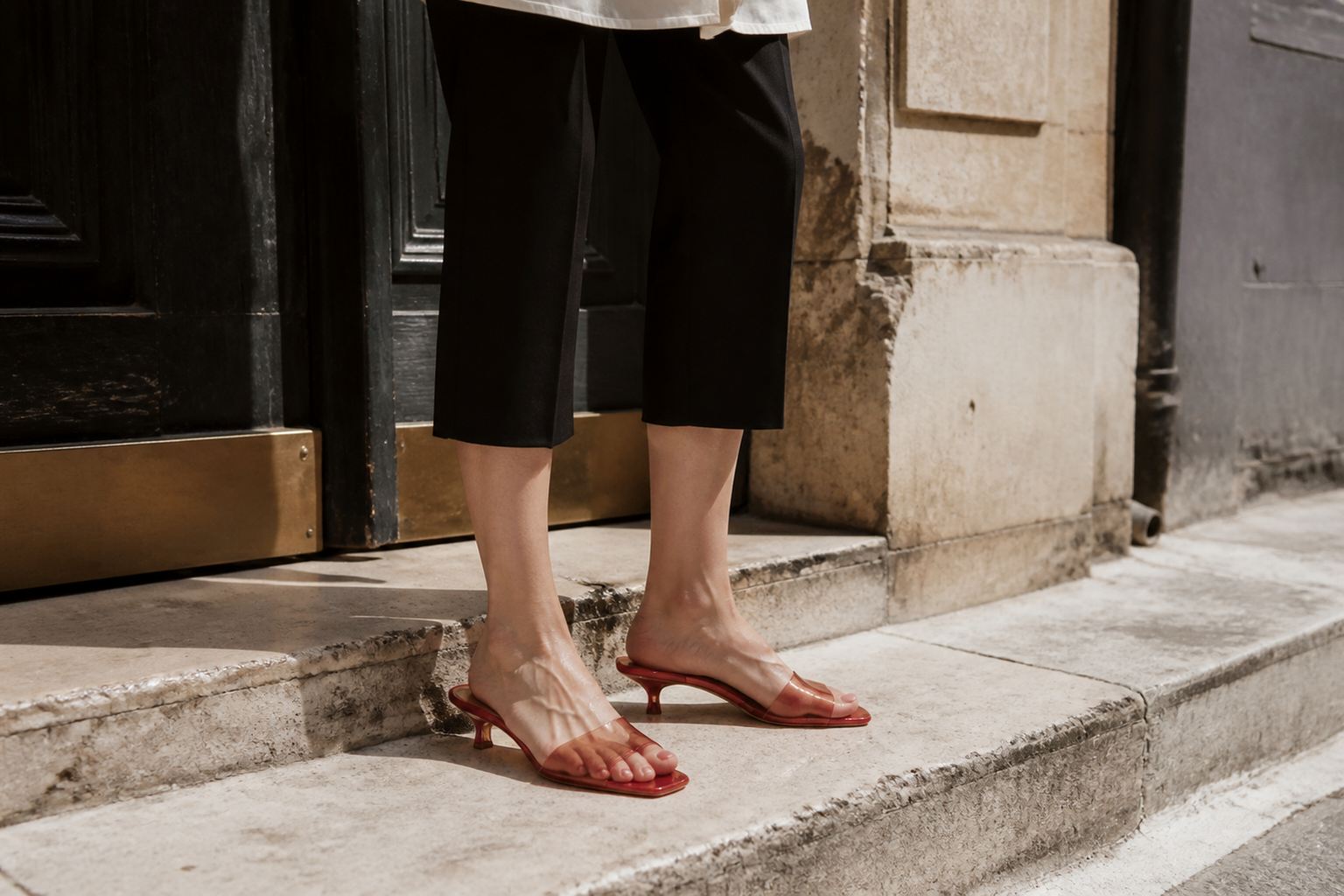 Faceless summer outfit with black cropped trousers and translucent red jelly kitten heels on worn limestone