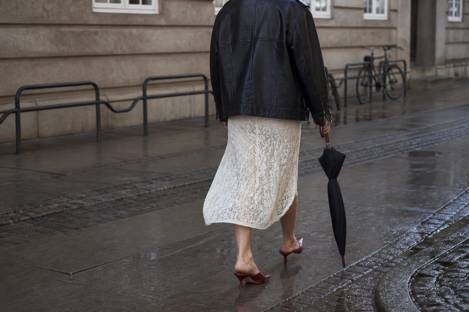 Faceless street-style look with a black leather jacket over a cream lace dress and dark red kitten heels on a wet Copenhagen street