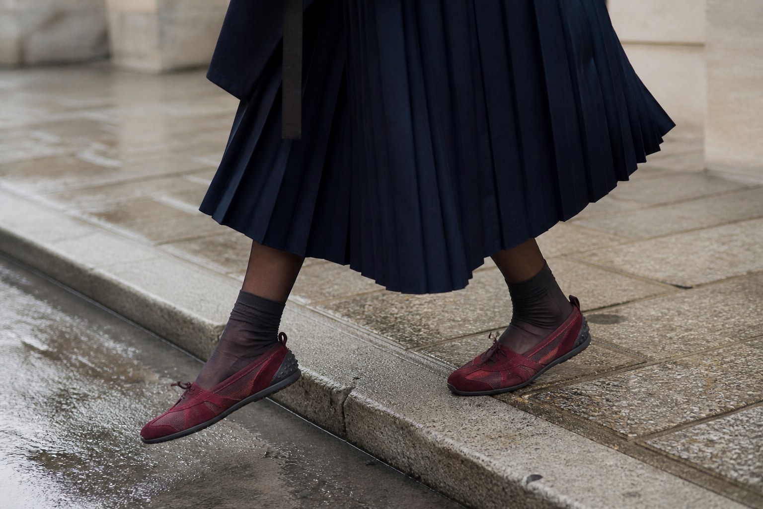 Burgundy technical runner flats with charcoal socks and a navy pleated skirt stepping from a wet city curb