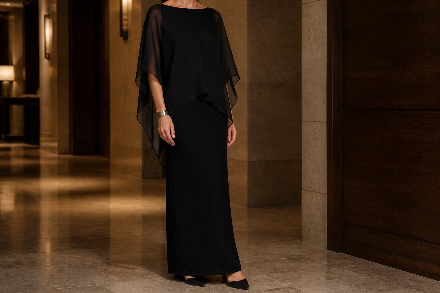 Black chiffon poncho layered over a matte black column dress in a warm stone hotel corridor