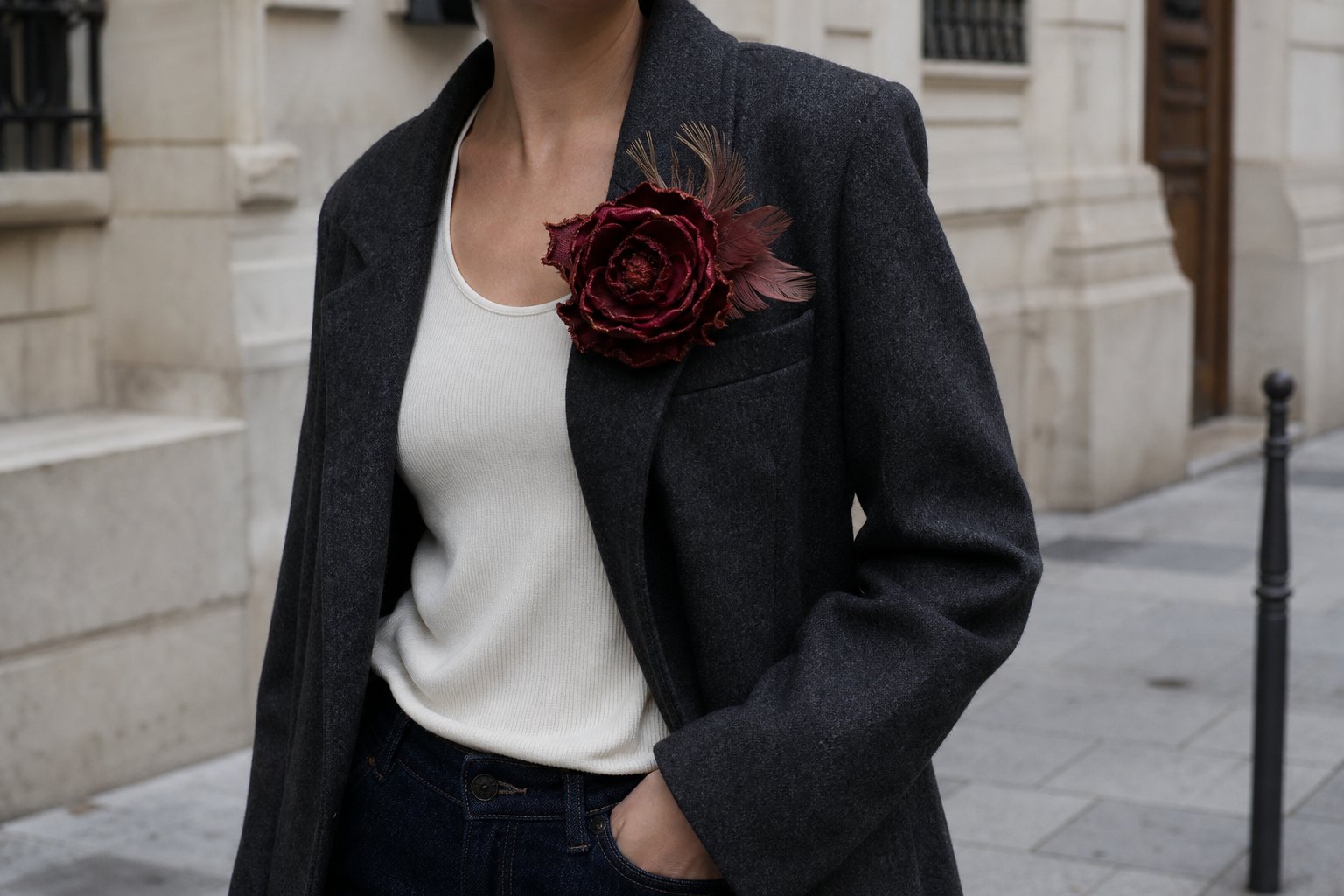 Large oxblood silk-and-feather brooch pinned high on a charcoal wool blazer, photographed from neck to waist on an overcast city street