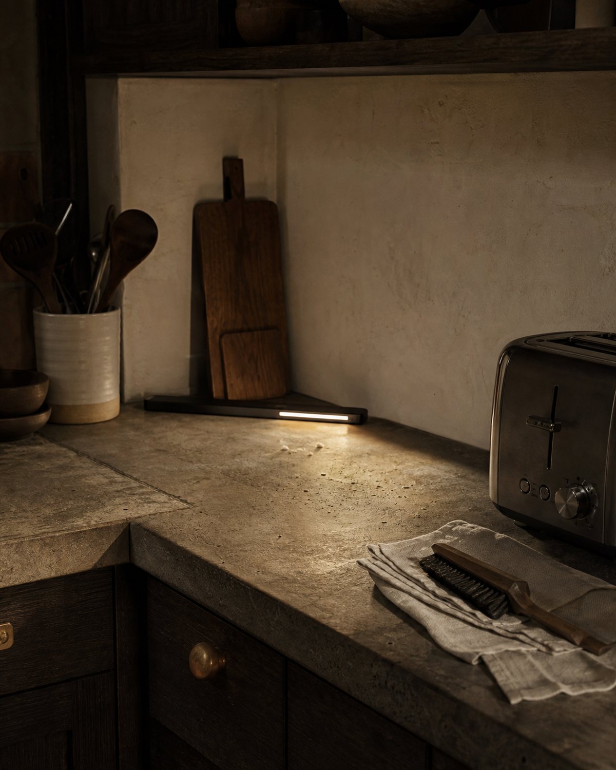 Low task light revealing a few crumbs on a dark stone kitchen counter beside a folded cloth and brush
