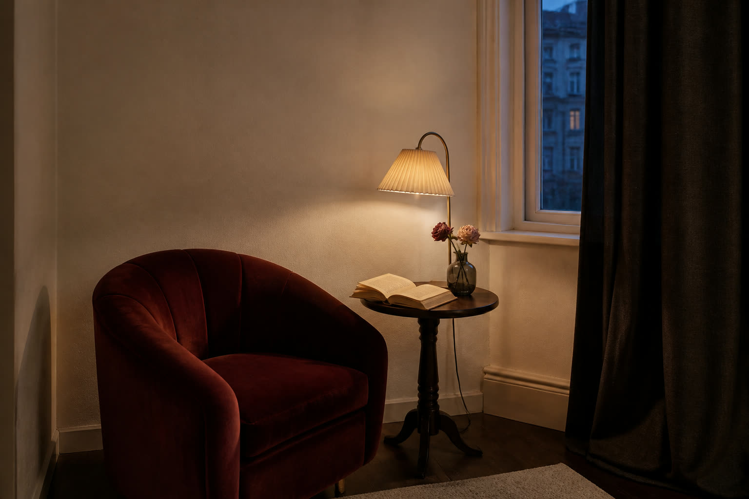 Compact reading corner with an oxblood velvet chair, pleated lamp, dark curtain, open book and two roses at dusk