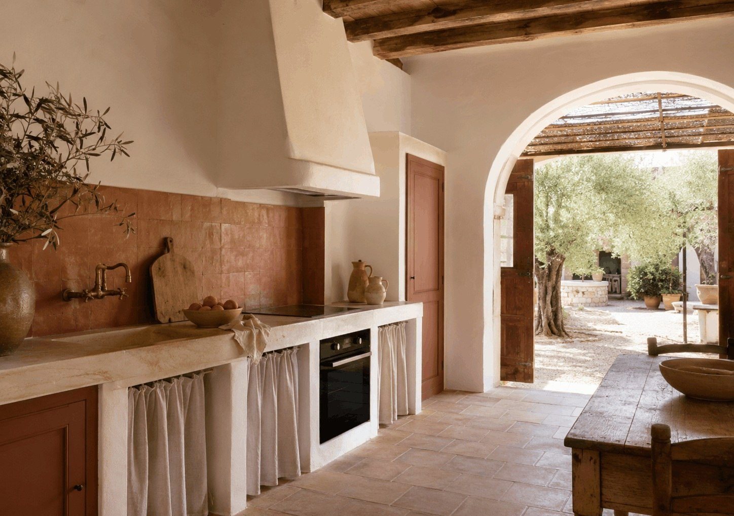 Terracotta and plaster kitchen opening to a sunlit courtyard with rustic timber table